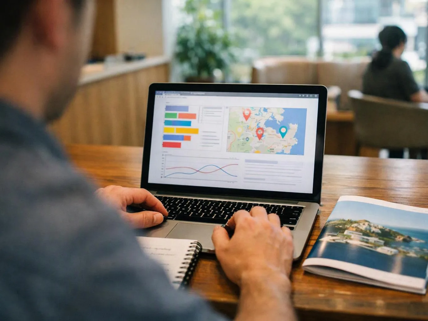 A person types on a laptop showing charts and a map, with a notebook and travel brochure on a wooden table