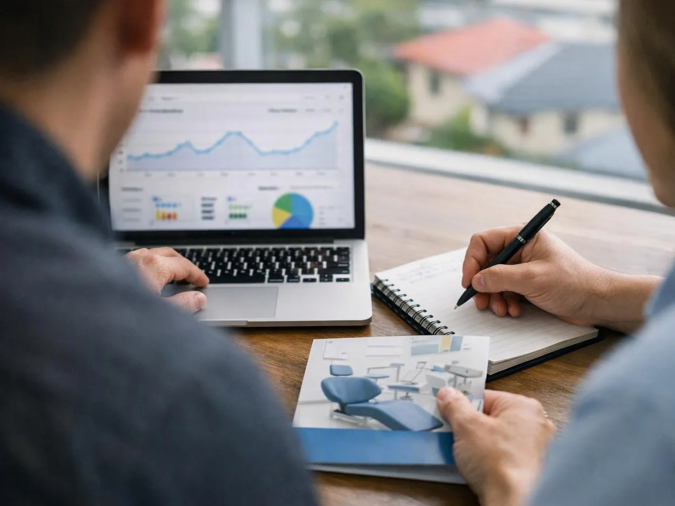 Two people review charts on a laptop and take notes at a table, with a brochure in hand